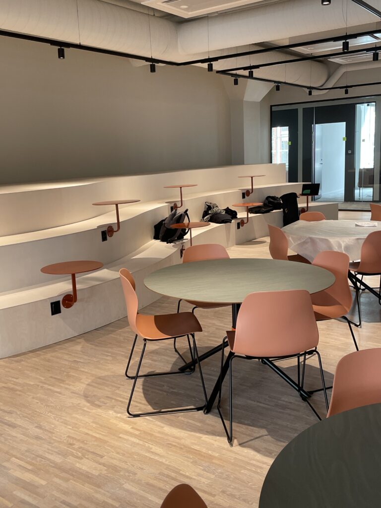 Pausyta med runda bord, terrakottafärgade stolar och trappad sittplatstribun i kontorsmiljö